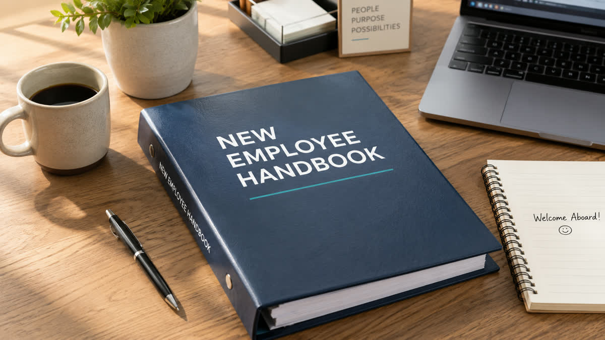 A binder titled New Employee Handbook sitting on a desk beside a coffee cup, a laptop, and a notebook that says Welcome Aboard.